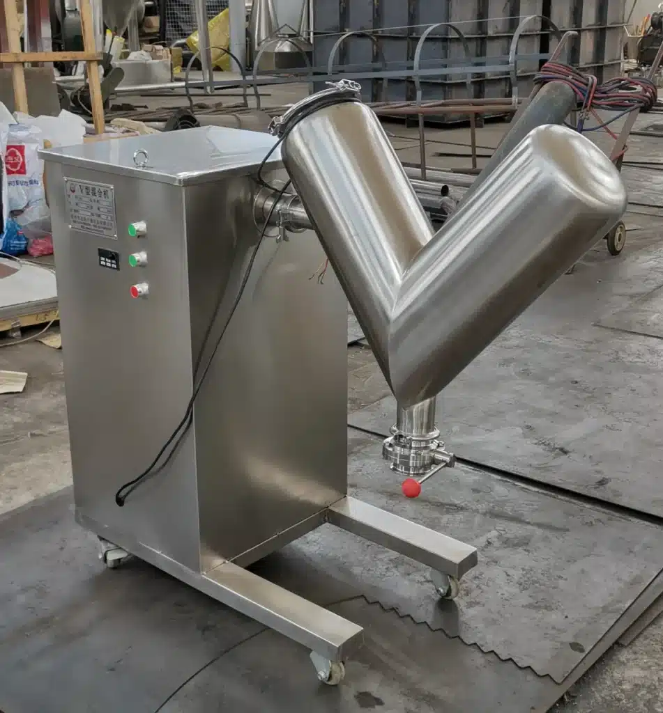 L'immagine mostra un grande miscelatore o mixer a V in acciaio inossidabile all'interno di quello che sembra essere uno stabilimento di produzione industriale o un'officina. Il miscelatore a V ha due camere di miscelazione cilindriche unite in una configurazione a V ed è montato su un telaio o supporto mobile in acciaio inossidabile. Sullo sfondo dello spazio di lavoro affollato si possono vedere vari tubi, manichette e altre attrezzature. V Blender