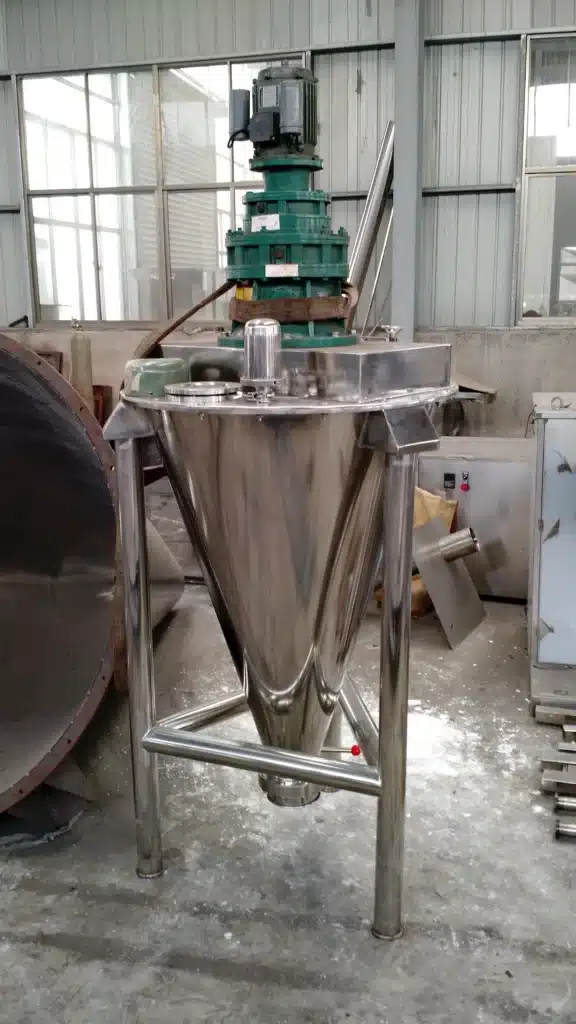 Miscelatore a coclea conica in acciaio inox in una fabbrica industriale L'immagine mostra un grande miscelatore o frullatore a coclea conica in acciaio inox situato in quello che sembra essere un impianto di produzione industriale o una fabbrica. Il miscelatore a coclea conica è costituito da una tramoggia o imbuto conico montato su un telaio o supporto in acciaio inox. I componenti chiave del miscelatore che si possono vedere includono il recipiente di miscelazione conico, l'albero e le pale dell'agitatore e l'uscita di scarico sul fondo del cono.