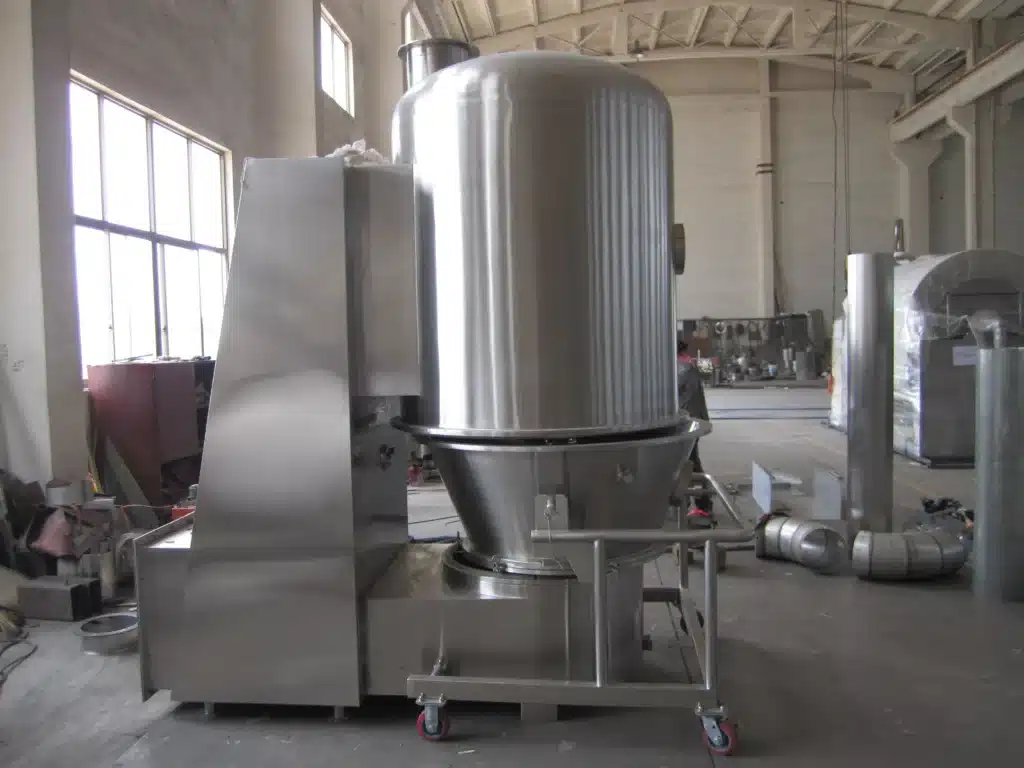 Большая вертикальная сушилка с псевдоожиженным слоем стоит в промышленном цеху. Высокий сосуд из нержавеющей стали с коническим основанием и центральной трубой на металлической раме с колесами предполагает мобильность и гибкость производственного пространства.