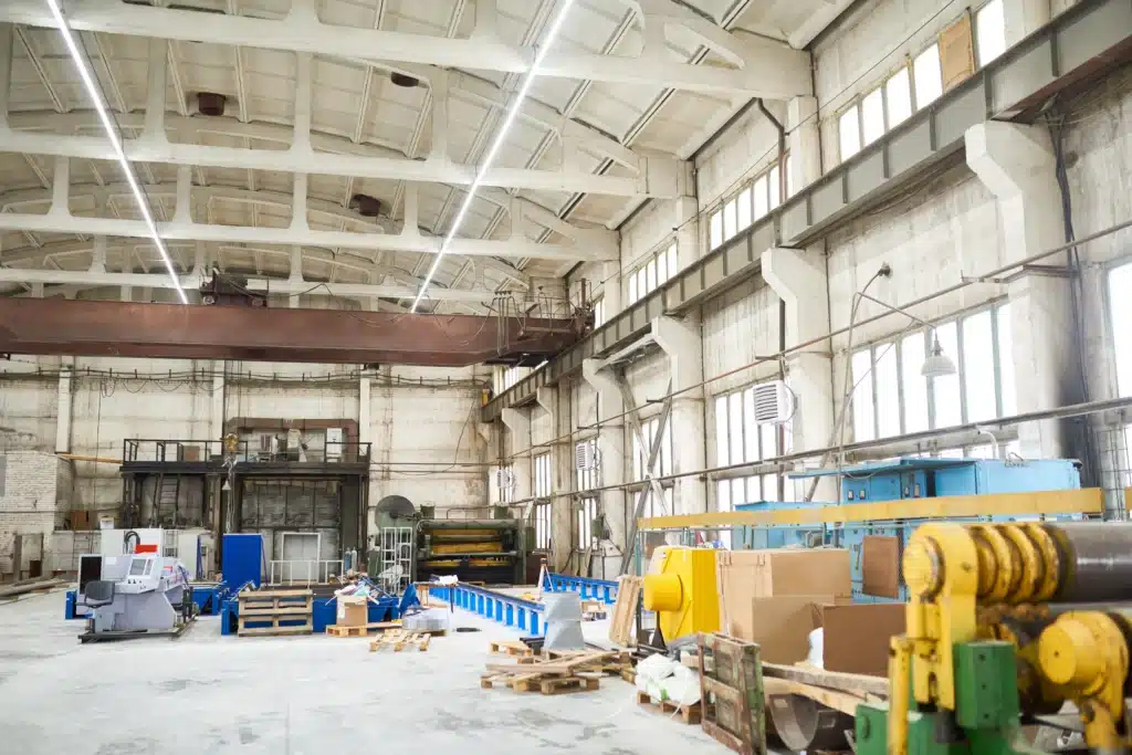 A spacious industrial warehouse with high ceilings, large windows, and various machinery and equipment scattered around. The space appears well-lit with overhead lights.