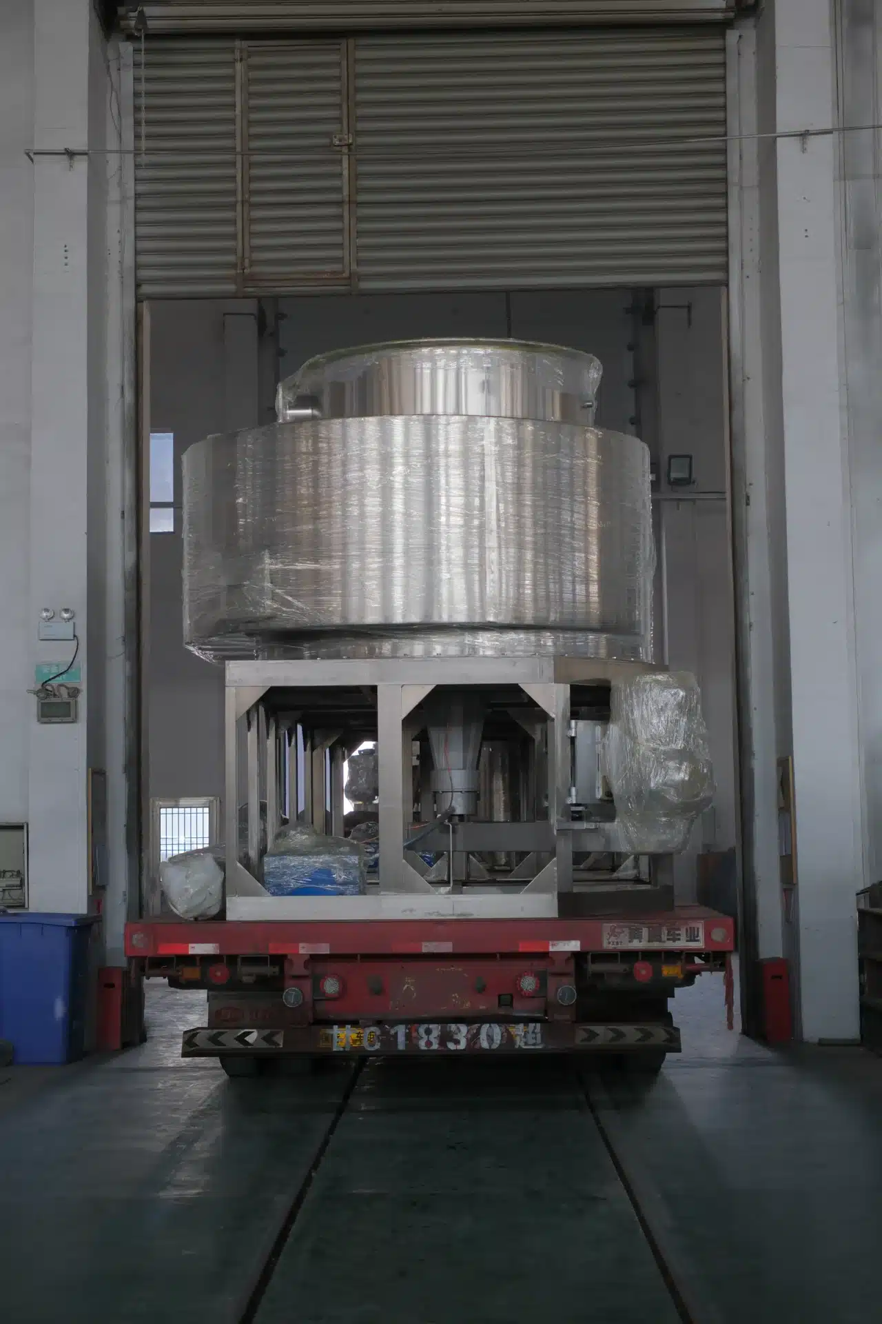 Máy sấy phun được bọc chắc chắn bằng vật liệu bảo vệ, được đưa lên xe tải sàn phẳng màu đỏ bên trong một cơ sở công nghiệp, sẵn sàng giao hàng. 